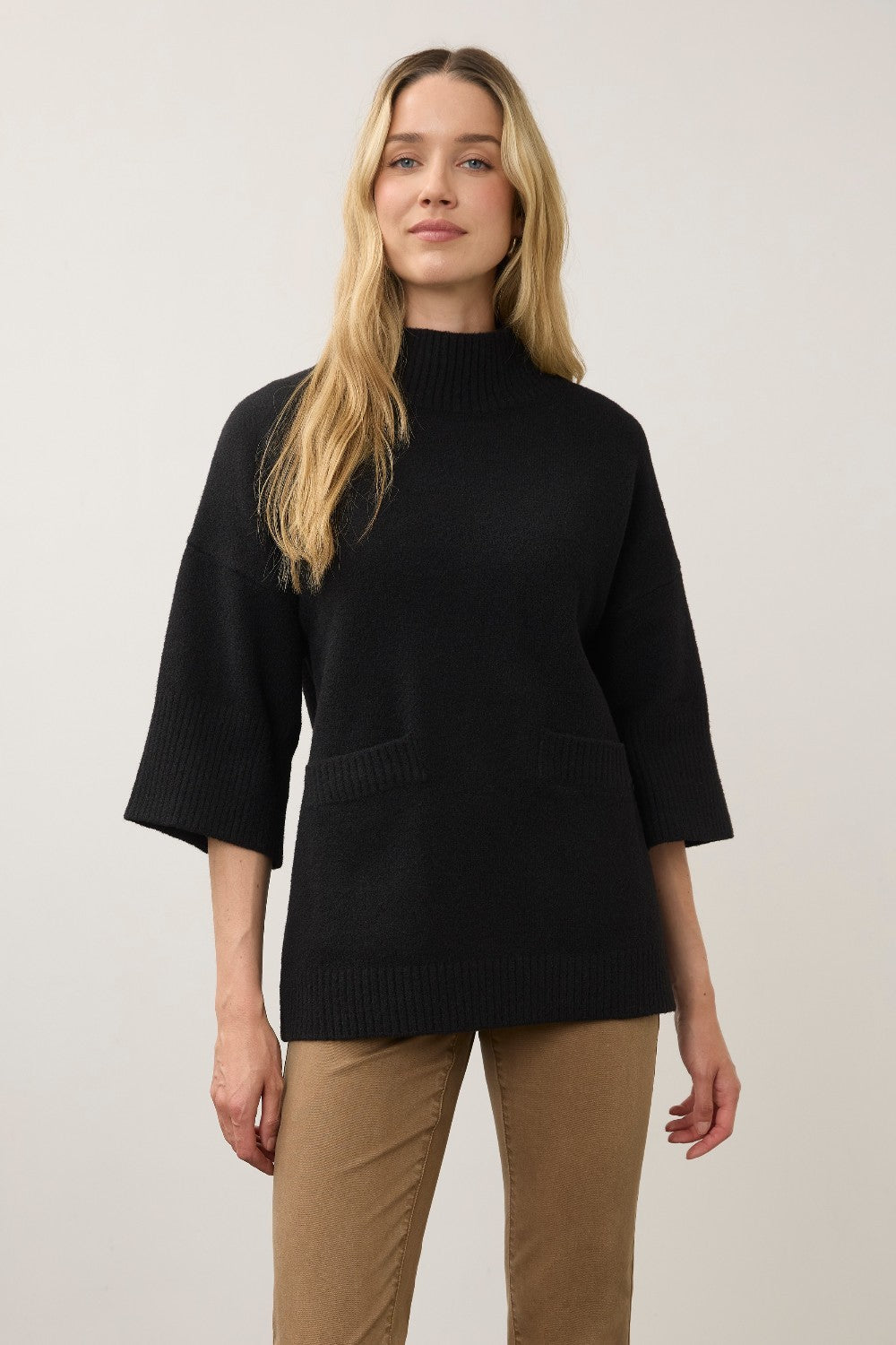 A woman modeling a black mock-neck knit sweater with three-quarter sleeves and front patch pockets, paired with tan pants, against a plain light gray studio background.
