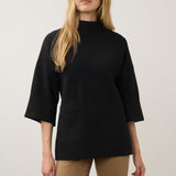 A woman modeling a black mock-neck knit sweater with three-quarter sleeves and front patch pockets, paired with tan pants, against a plain light gray studio background.