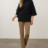 A woman modeling a black mock-neck knit top with wide three-quarter sleeves, paired with tan trousers and black ankle boots, standing against a plain off-white wall.