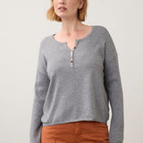 Woman wearing a grey sweater with multi coloured button detail at neck line with orange pants on a light background.