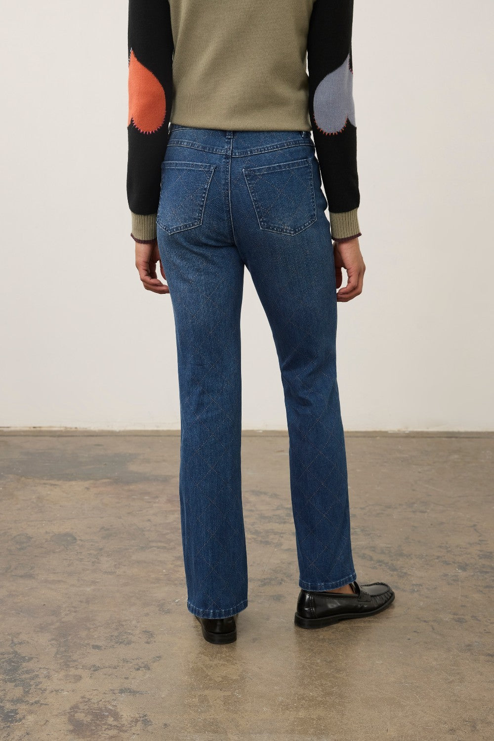 A person shown from the back wearing blue straight-leg jeans, a beige knit sweater with black, orange, and gray color-blocked sleeves, and black loafers, standing against a plain off-white wall and concrete floor.