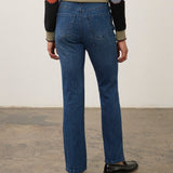 A person shown from the back wearing blue straight-leg jeans, a beige knit sweater with black, orange, and gray color-blocked sleeves, and black loafers, standing against a plain off-white wall and concrete floor.