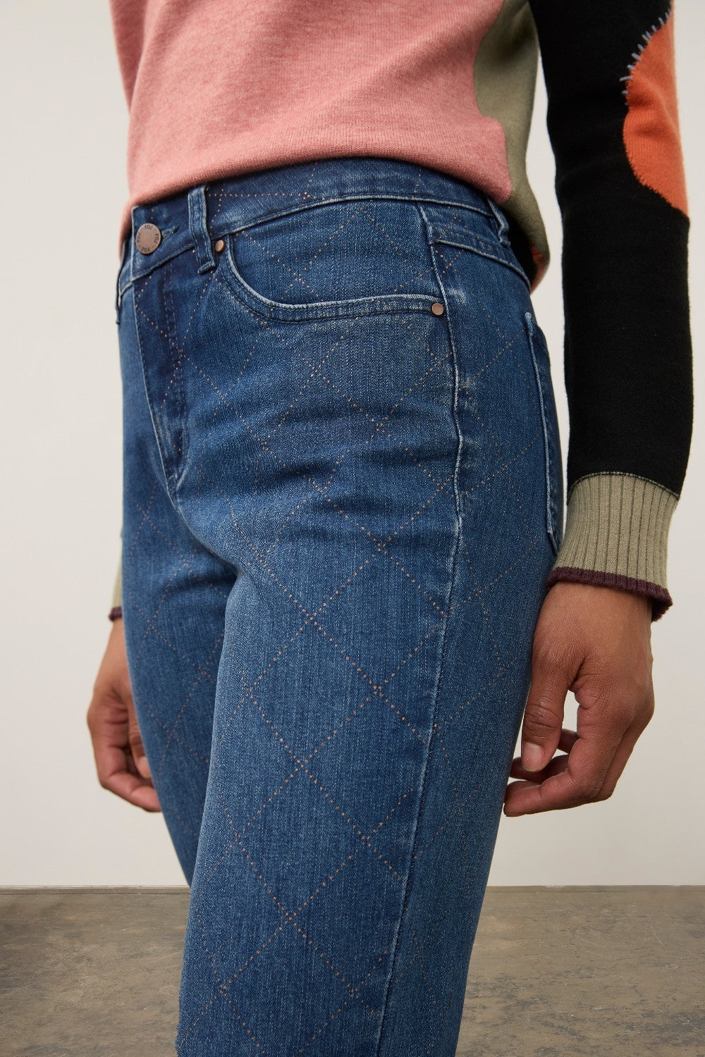 Close-up of a person wearing medium-wash blue jeans with a subtle diamond argyle knit pattern on the front, paired with a pink and black color-blocked sweater.