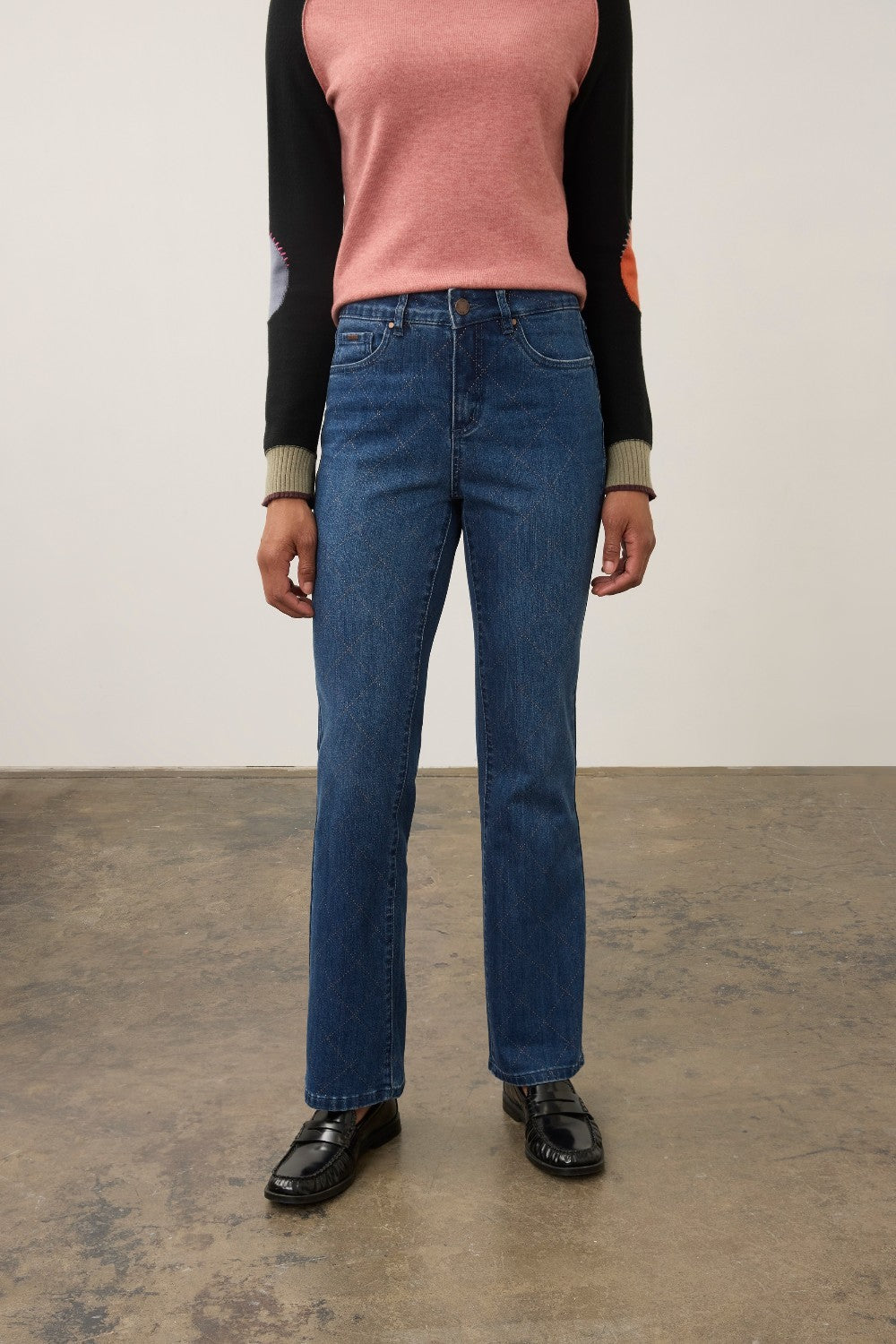 A model wearing a pair of straight-leg blue jeans, paired with a pink and black long-sleeve top and black leather loafers, standing against a neutral studio background.