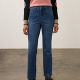 A model wearing a pair of straight-leg blue jeans, paired with a pink and black long-sleeve top and black leather loafers, standing against a neutral studio background.