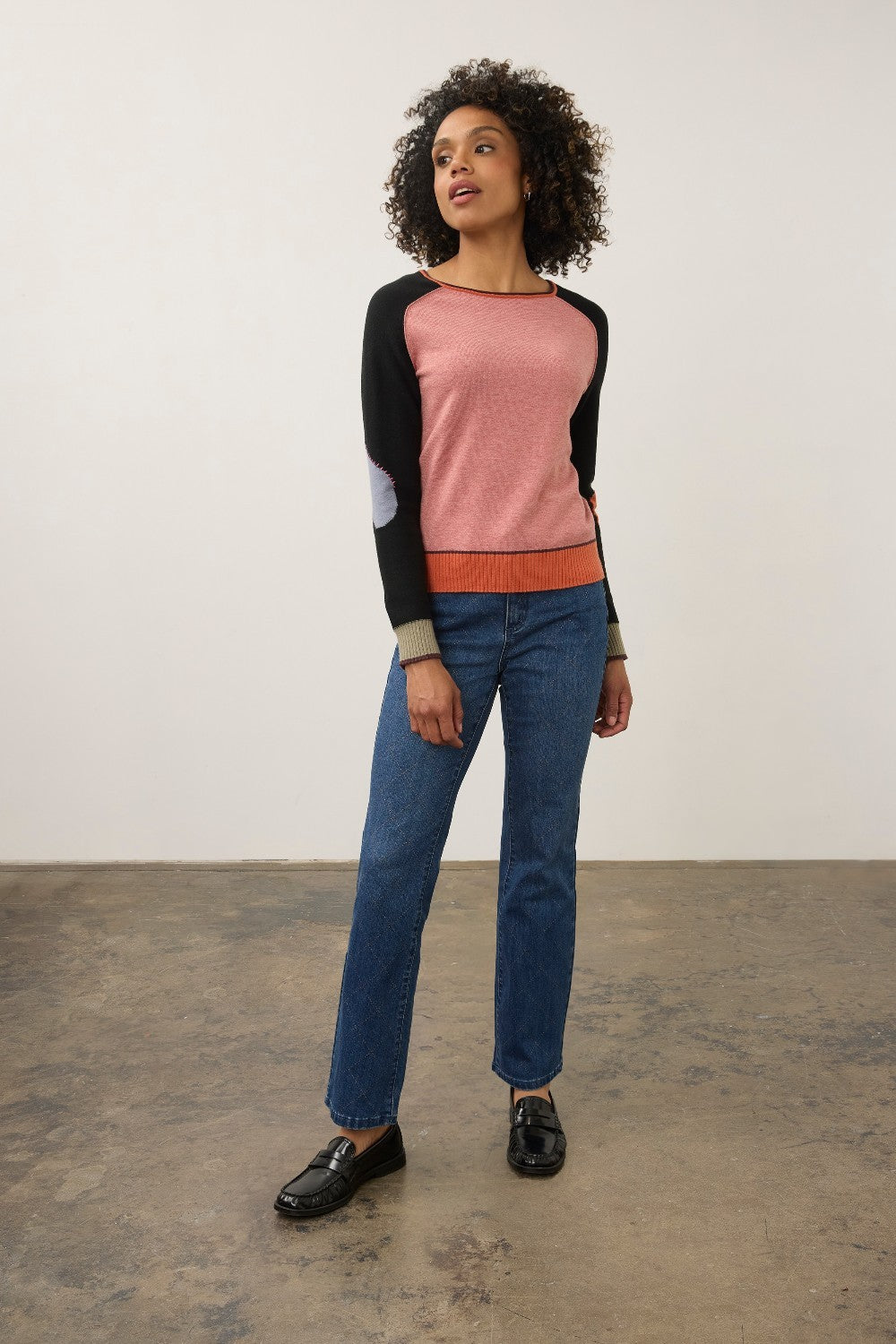 A woman modeling a colorblock raglan sweater with blue jeans and black loafers, standing against a plain off-white wall and concrete floor.