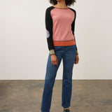A woman modeling a colorblock raglan sweater with blue jeans and black loafers, standing against a plain off-white wall and concrete floor.