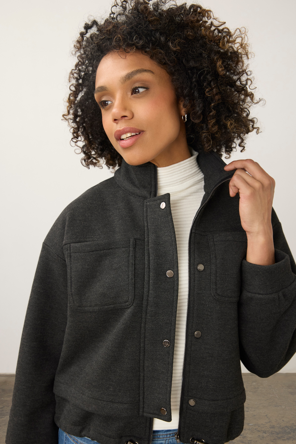 A woman with curly dark hair wearing a dark charcoal gray cropped button-up jacket over a white ribbed turtleneck sweater and blue jeans, photographed against a neutral light gray studio background.