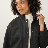 A woman with curly dark hair wearing a dark charcoal gray cropped button-up jacket over a white ribbed turtleneck sweater and blue jeans, photographed against a neutral light gray studio background.