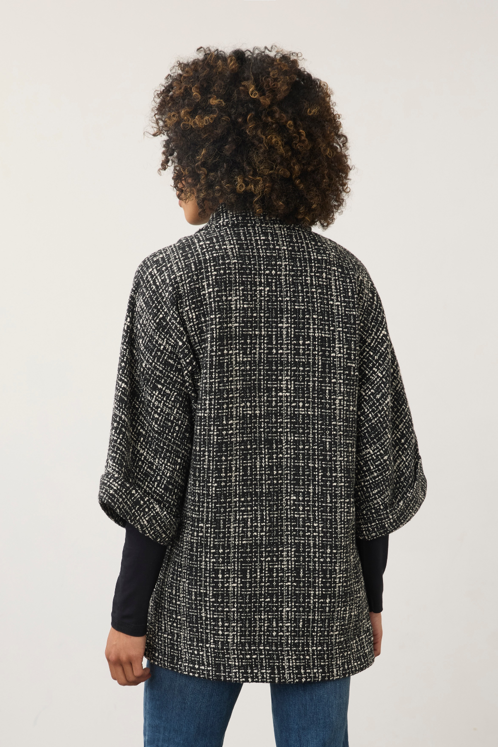 A person shown from the back wearing a black and white textured tweed cape jacket over a black long-sleeve top and blue jeans, against a plain light gray background.