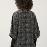 A person shown from the back wearing a black and white textured tweed cape jacket over a black long-sleeve top and blue jeans, against a plain light gray background.