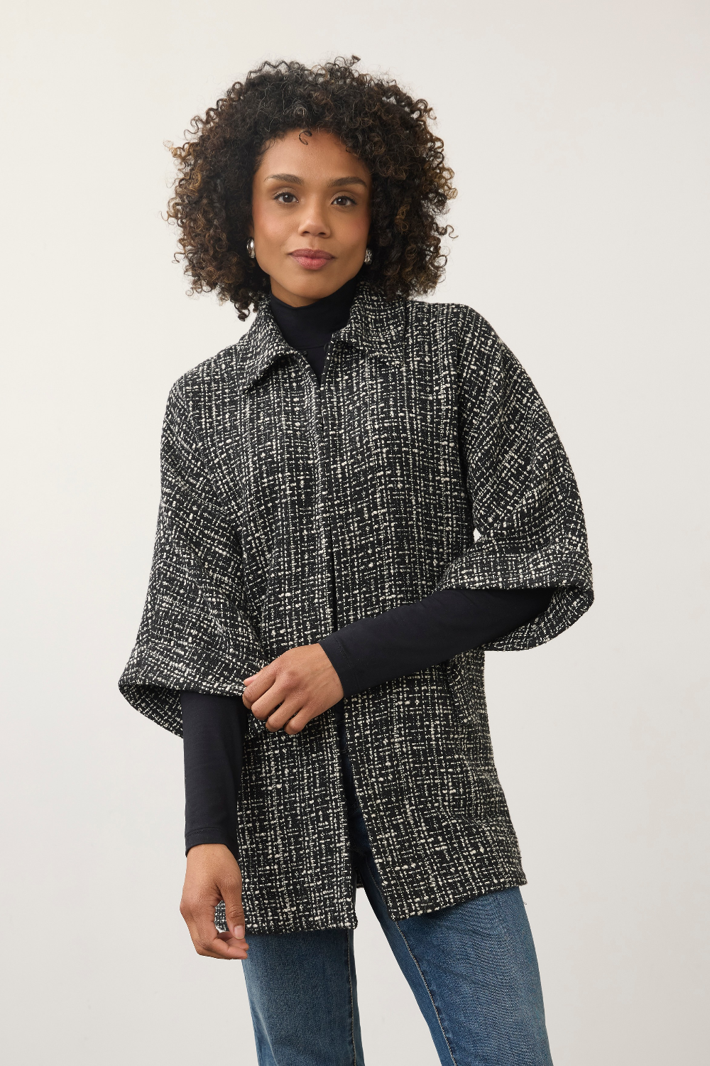 A woman with curly dark hair wearing a black and white textured tweed cape jacket over a black turtleneck and blue jeans, standing against a plain light gray studio background.