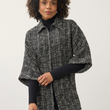 A woman with curly dark hair wearing a black and white textured tweed cape jacket over a black turtleneck and blue jeans, standing against a plain light gray studio background.