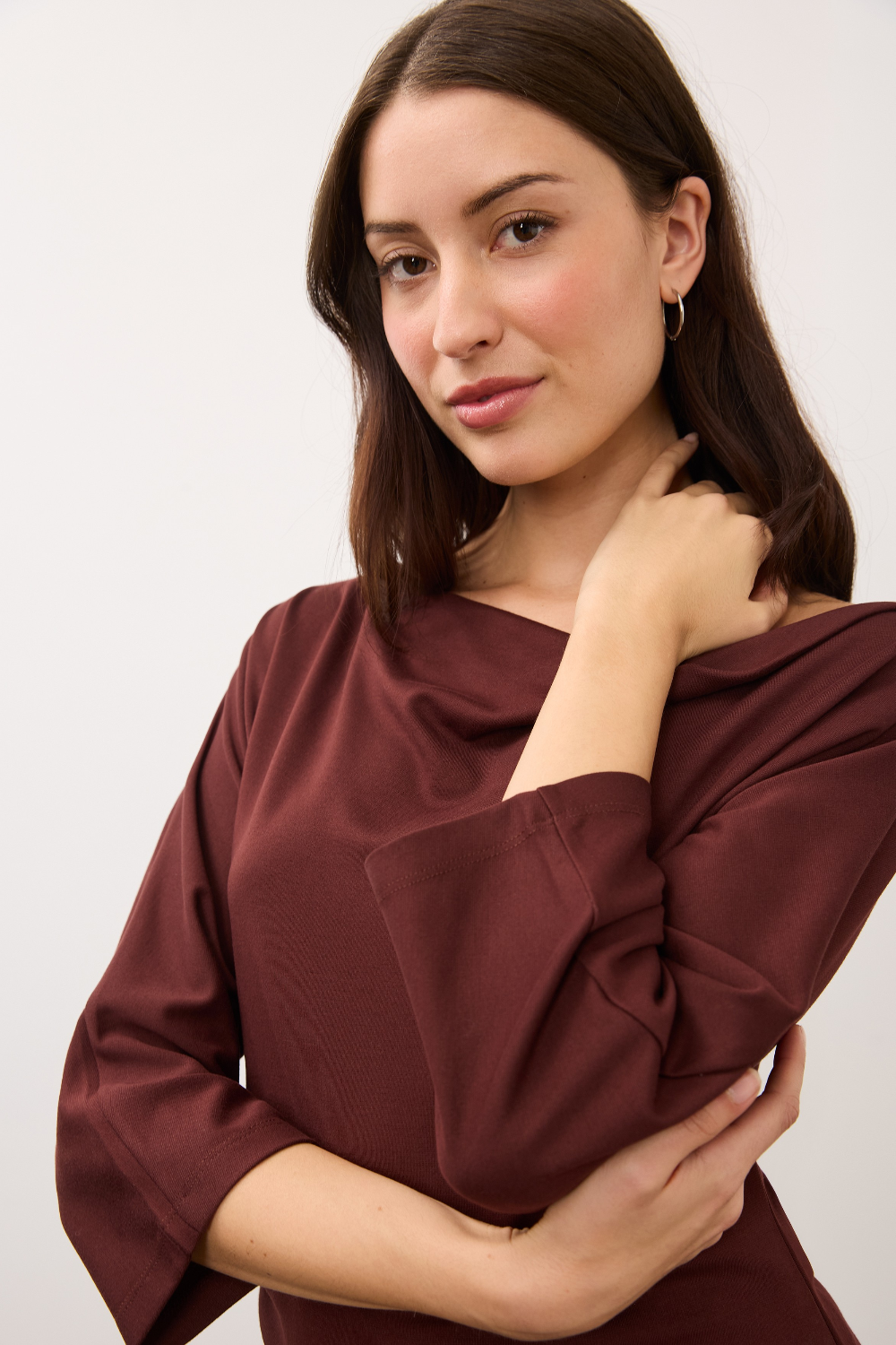 A woman modeling a draped, three-quarter sleeve top in a deep burgundy color against a plain light gray background.
