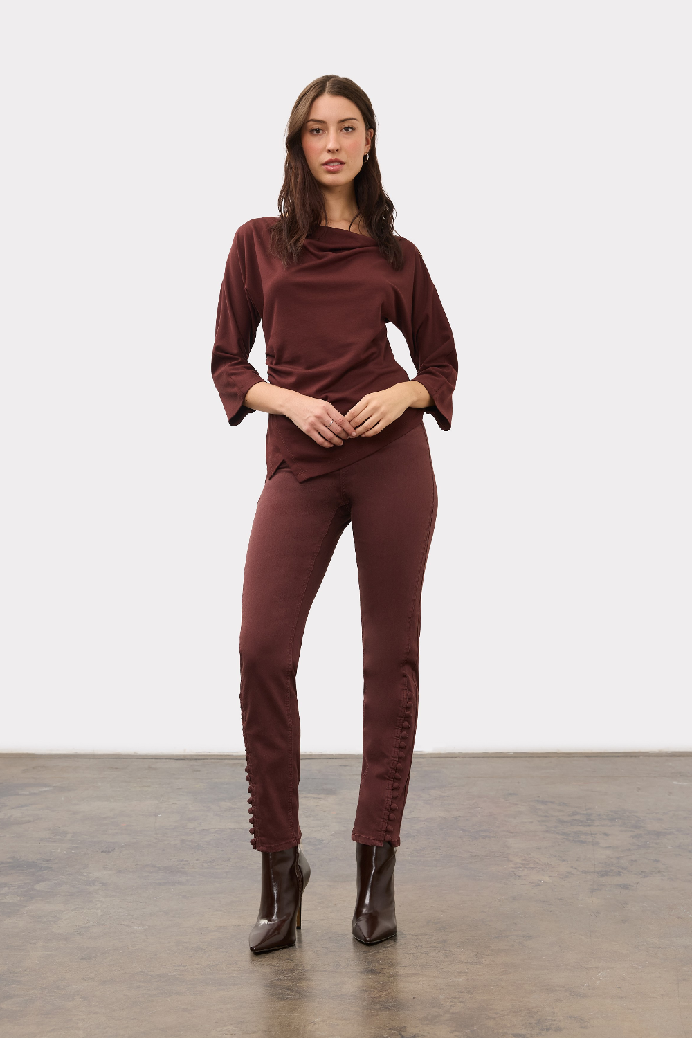A woman modeling a draped, three-quarter sleeve top in a deep burgundy color, paired with matching pants and boots, against a plain light gray background.
