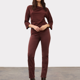 A woman modeling a draped, three-quarter sleeve top in a deep burgundy color, paired with matching pants and boots, against a plain light gray background.