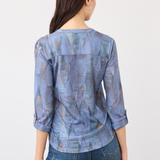 Woman, shown from the back, wearing a blue mixed pattern 3/4 sleeve top with blue jeans on a light background.
