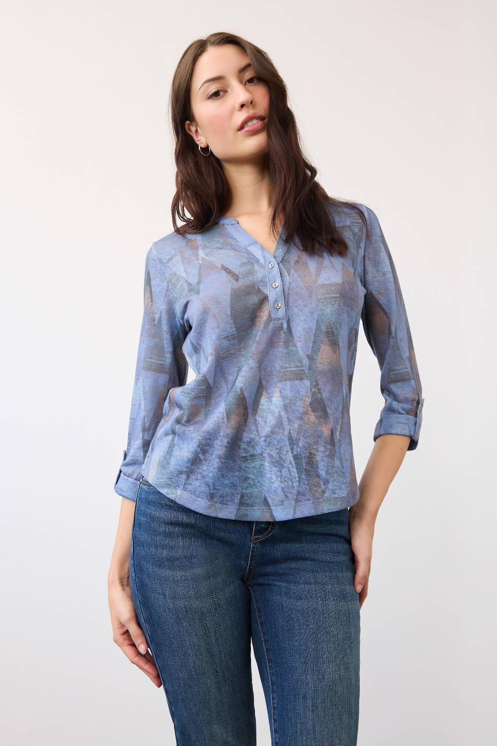 Woman wearing a blue mixed pattern 3/4 sleeve henley top with blue jeans on a light background.