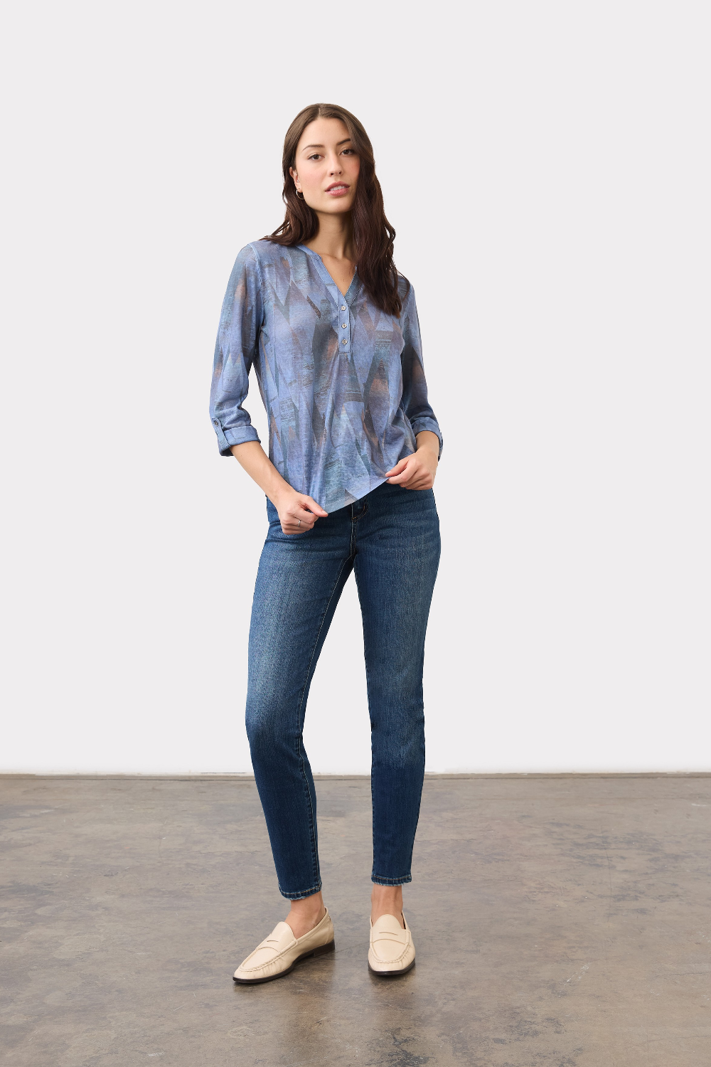 Woman wearing a blue mixed pattern 3/4 sleeve henley top with blue jeans and tan loafers on a light background.