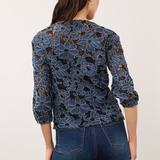 Woman, shown from the back, wearing a blue and black floral lace 3/4 sleeve v-neck top with blue jeans on a light background.