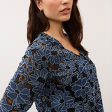 Woman wearing a blue and black floral lace 3/4 sleeve v-neck top on a light background.