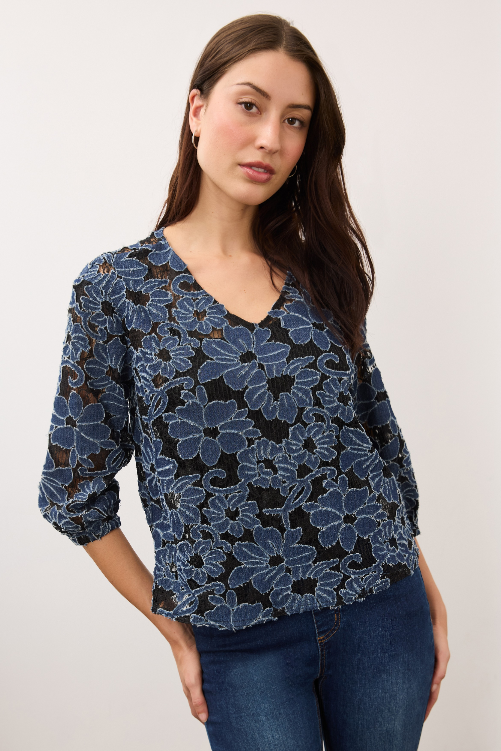 Woman wearing a blue and black floral lace 3/4 sleeve v-neck top with blue jeans on a light background.