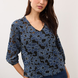 Woman wearing a blue and black floral lace 3/4 sleeve v-neck top with blue jeans on a light background.