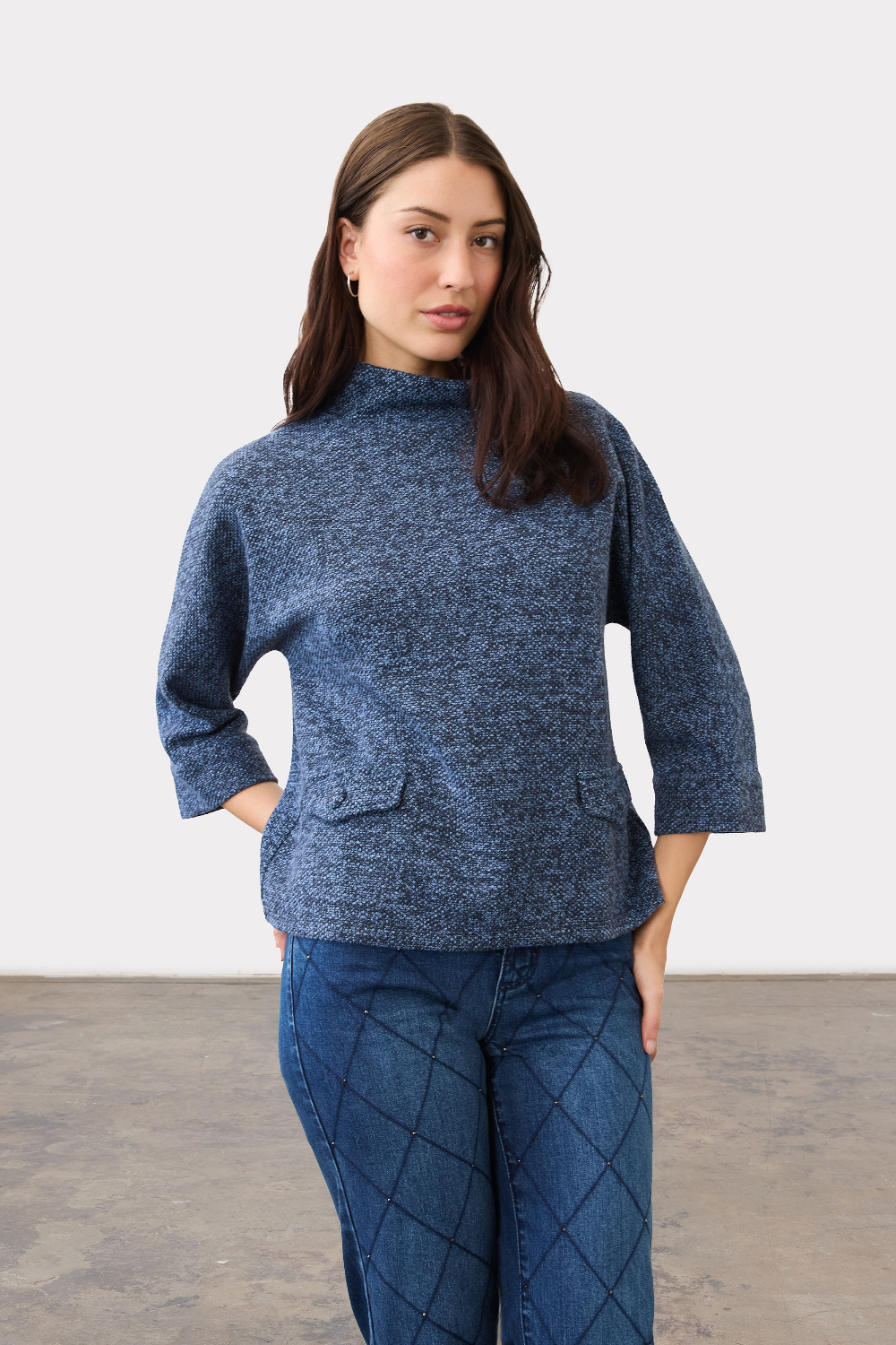 Woman wearing a blue 3/4 sleeve mock neck top, with pocket flap detailing, with blue jeans on a neutral backdrop.
