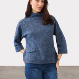 Woman wearing a blue 3/4 sleeve mock neck top, with pocket flap detailing, with blue jeans on a neutral backdrop.