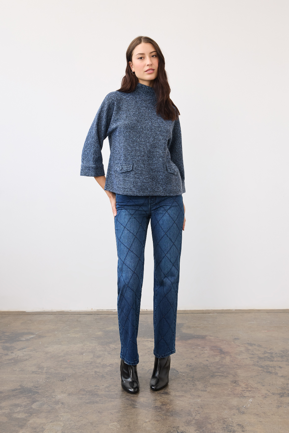 Woman wearing a blue 3/4 sleeve mock neck top, with pocket flap detailing, with blue jeans and black boots on a neutral backdrop.