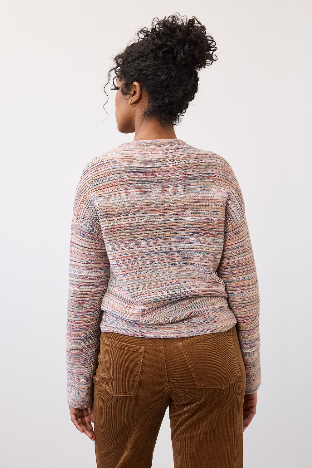 Woman, shown from the back, wearing a multi coloured striped sweater with brown cords on a light background.