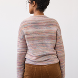 Woman, shown from the back, wearing a multi coloured striped sweater with brown cords on a light background.