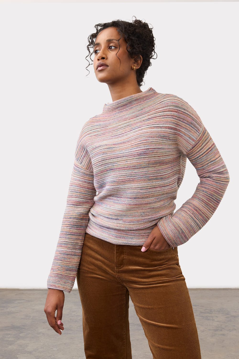 Woman wearing a multi coloured striped sweater with brown cords on a light background.