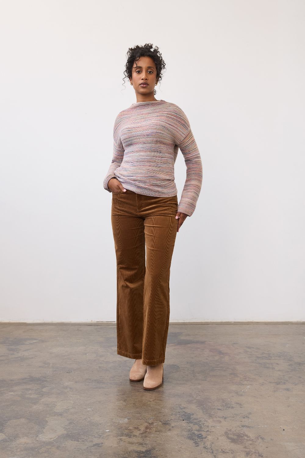 Woman wearing a multi coloured striped sweater with brown cords and tan boots on a light background.