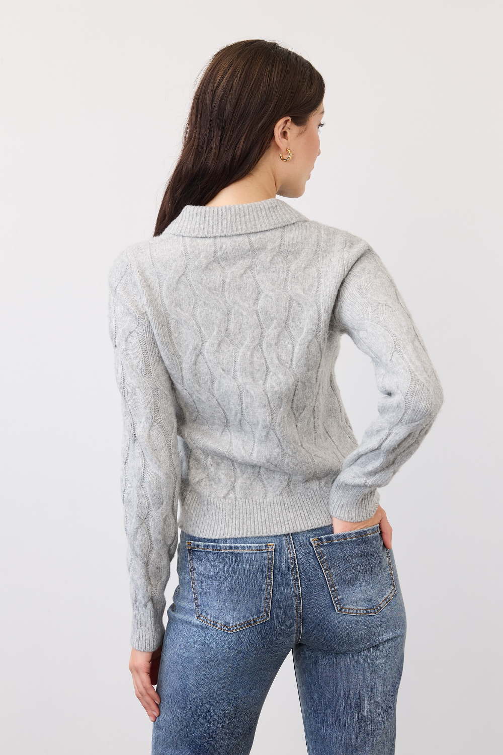 Woman, shown from the back, wearing a gray cable knit sweater, paired with blue jeans, photographed in front of a light studio backdrop.