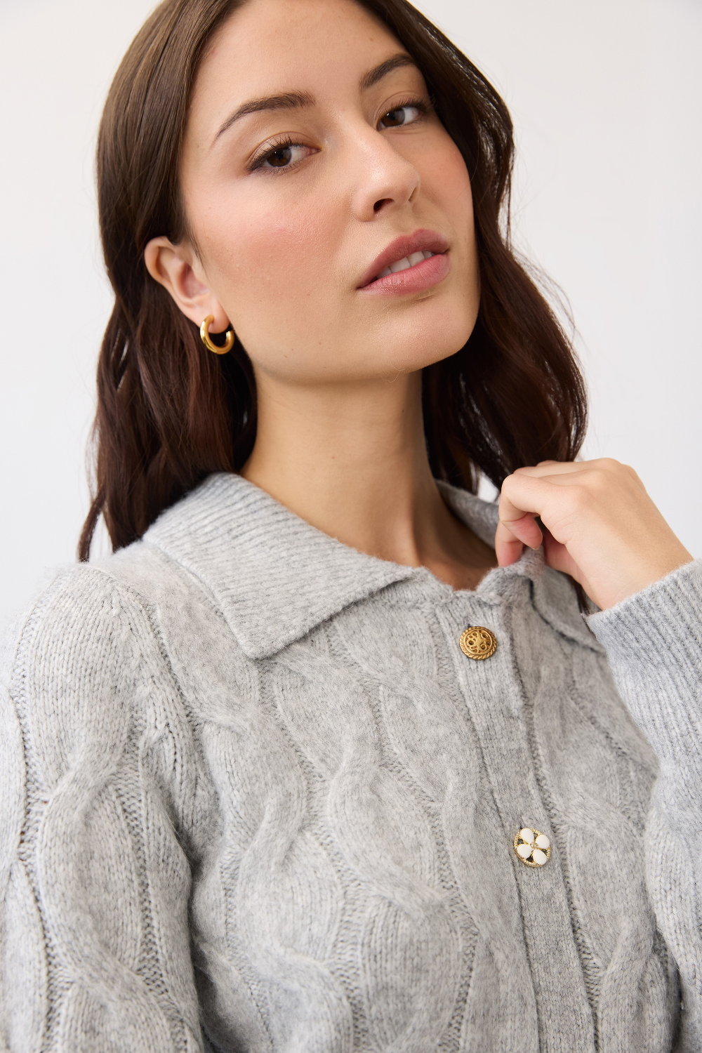 Close up of a woman wearing a gray cable knit cardigan, with a collar and fancy buttons, photographed in front of a light studio backdrop.