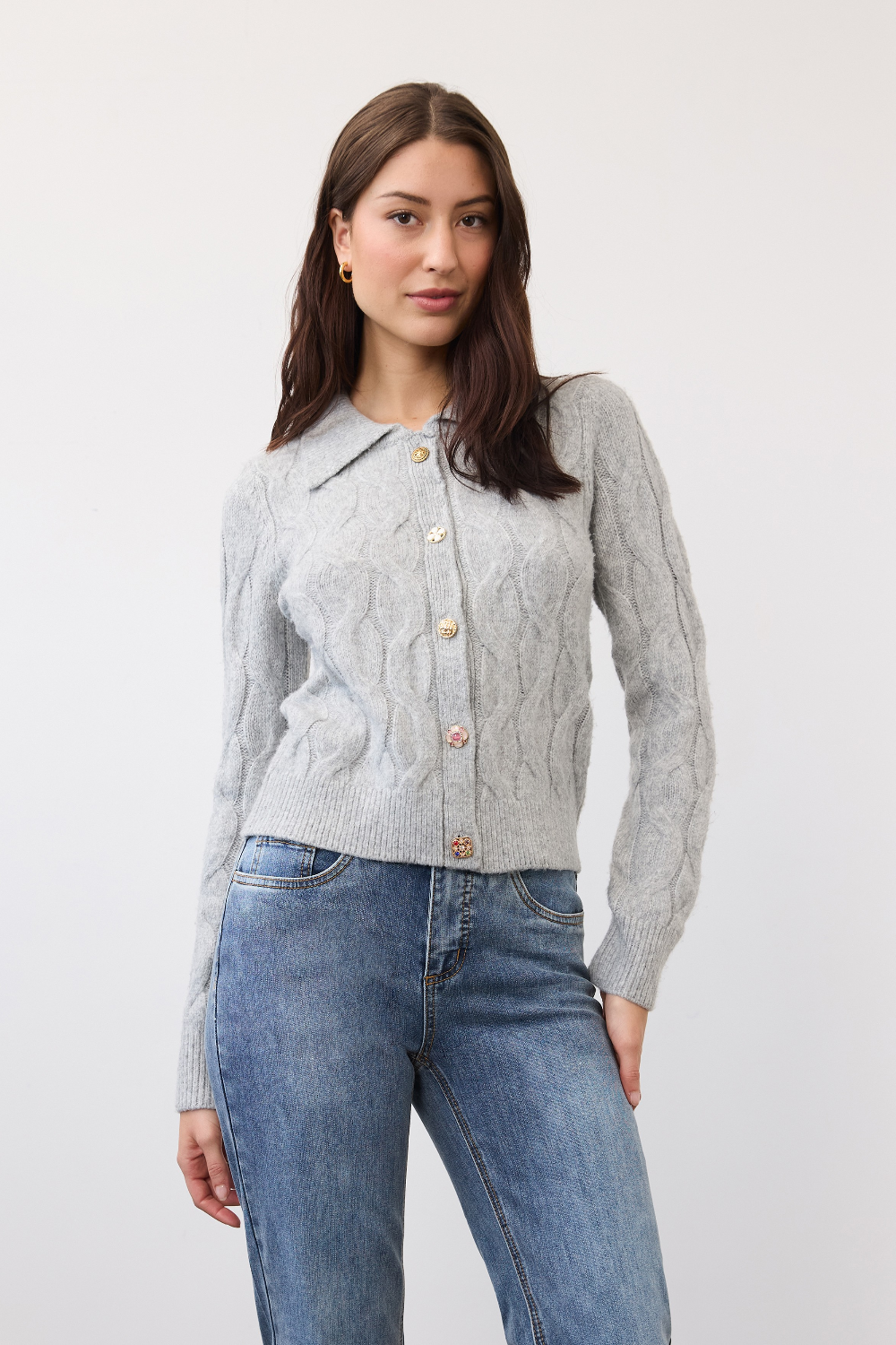 Woman wearing a gray cable knit cardigan, with a collar and fancy buttons, paired with blue jeans, photographed in front of a light studio backdrop.