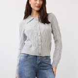 Woman wearing a gray cable knit cardigan, with a collar and fancy buttons, paired with blue jeans, photographed in front of a light studio backdrop.