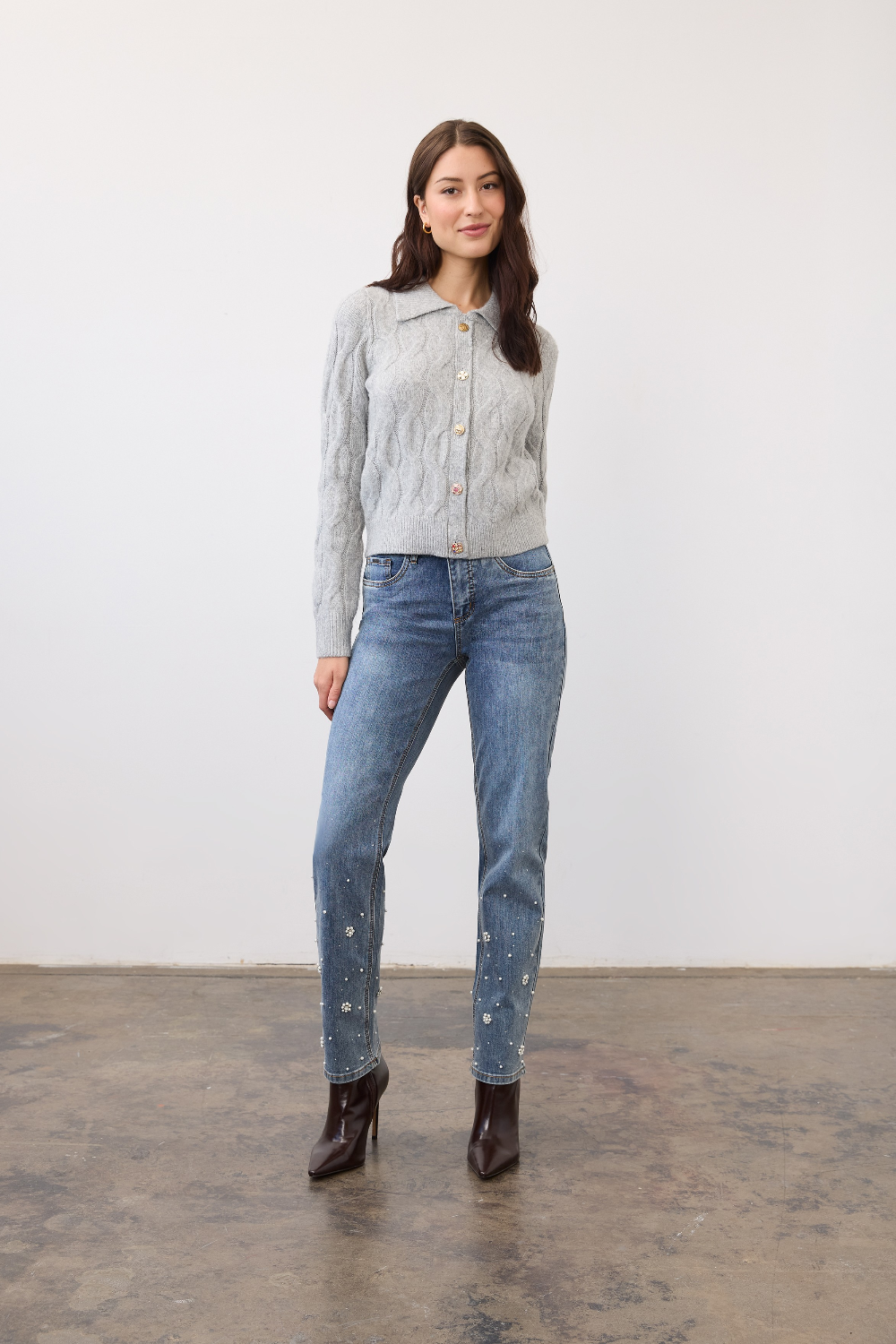 Woman wearing a gray cable knit cardigan, with a collar and fancy buttons, paired with blue jeans and burgundy boots, photographed in front of a light studio backdrop.