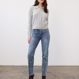 Woman wearing a gray cable knit cardigan, with a collar and fancy buttons, paired with blue jeans and burgundy boots, photographed in front of a light studio backdrop.