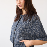 A woman wearing a navy blue and white speckled knit poncho-style cardigan with front pockets, shown from the waist up against a plain light background.