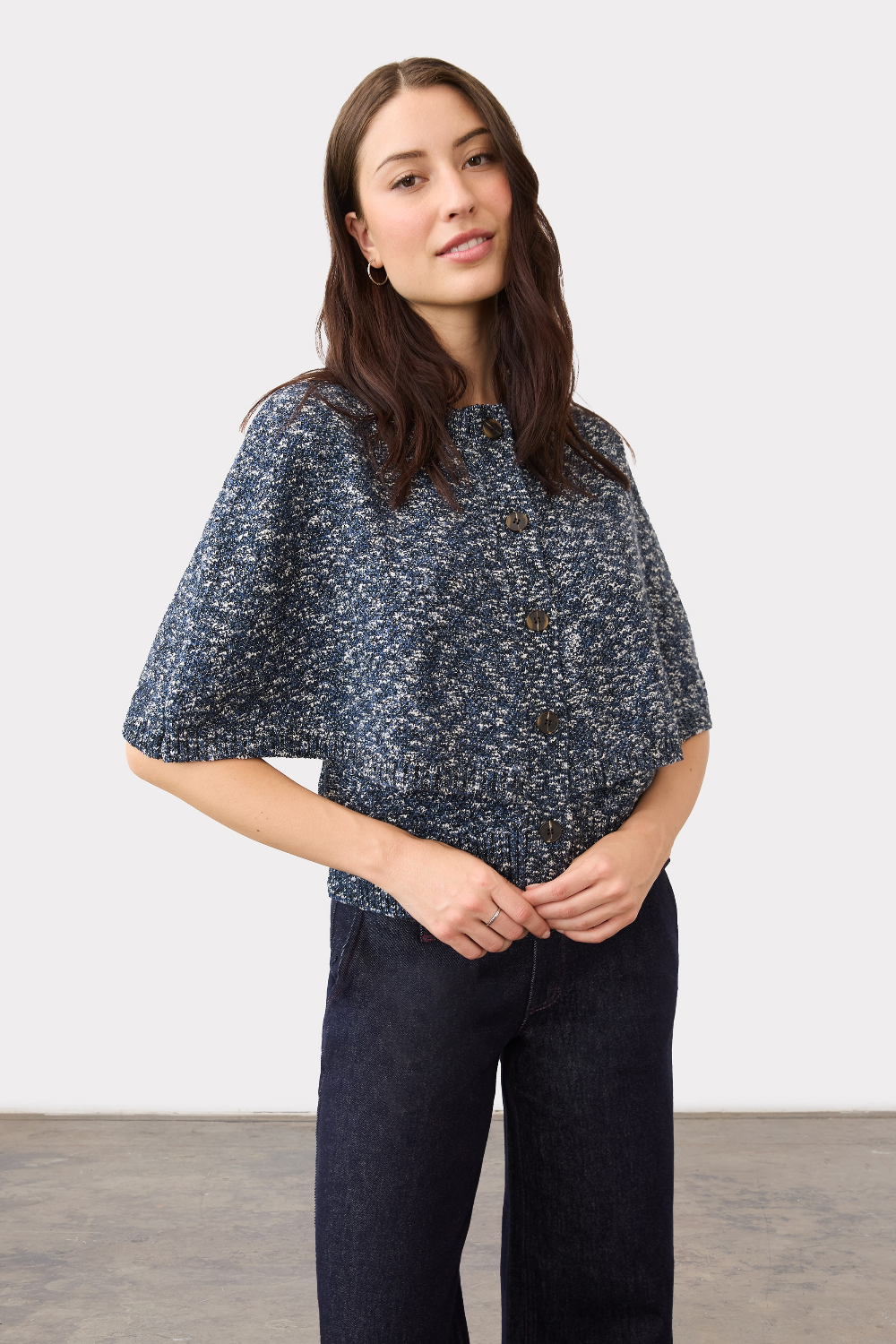 A woman modeling a short-sleeve button-front sweater with a blue and white speckled pattern, paired with dark jeans, against a plain light gray studio background.
