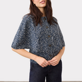 A woman modeling a short-sleeve button-front sweater with a blue and white speckled pattern, paired with dark jeans, against a plain light gray studio background.