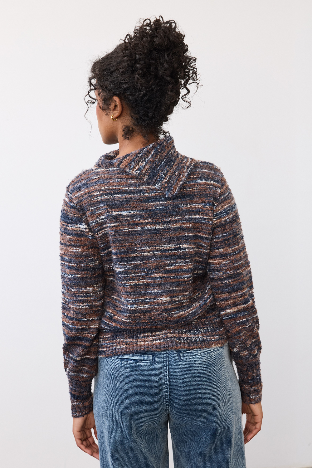 Woman, shown from the back, wearing a multi coloured striped sweater with blue cords on a light background. 
