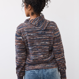 Woman, shown from the back, wearing a multi coloured striped sweater with blue cords on a light background. 