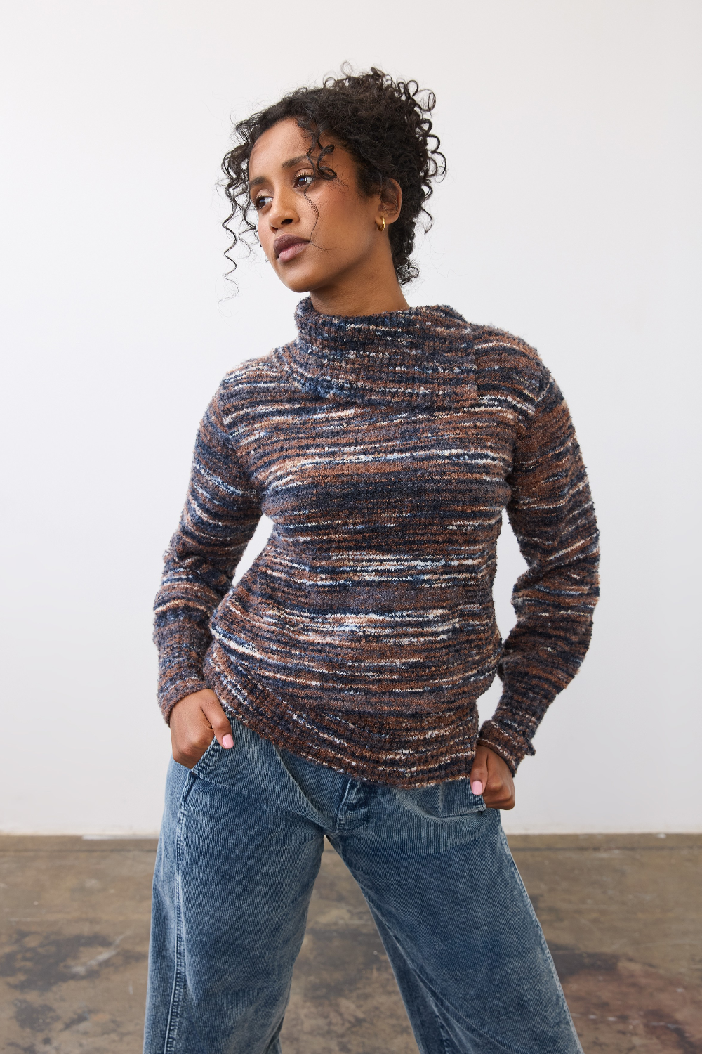 Woman wearing a multi coloured striped sweater with blue cords on a light background.