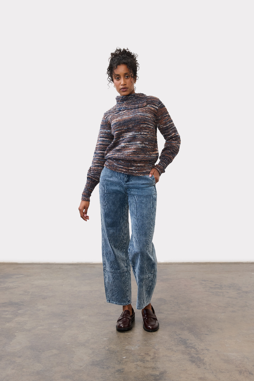 Woman wearing a multi coloured striped sweater with blue cords and brown loafers on a light background.