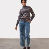 Woman wearing a multi coloured striped sweater with blue cords and brown loafers on a light background.