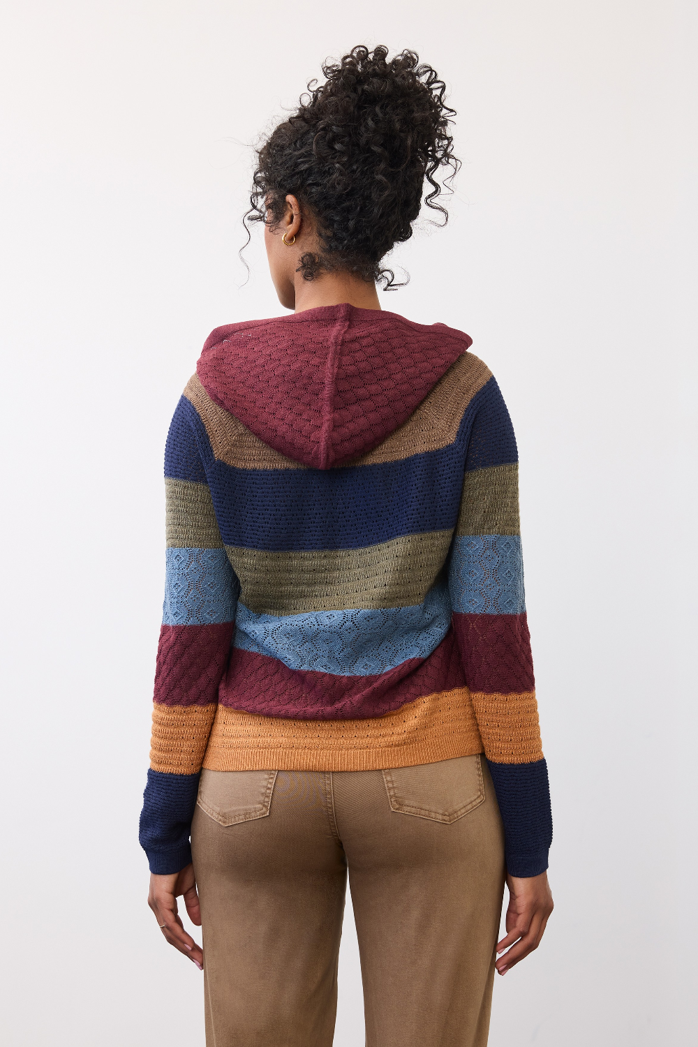 Woman, shown from behind, wearing a multi coloured striped knit hoodie with tan pants against a light backdrop.