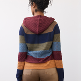 Woman, shown from behind, wearing a multi coloured striped knit hoodie with tan pants against a light backdrop.